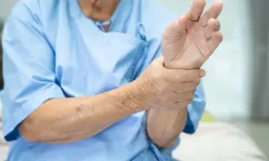 Main d'une personne âgée en blouse bleue, tenant son poignet, peut-être un pouce qui bouge tout seul.