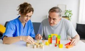 Une aide-soignante en blouse bleue joue avec des cubes en bois avec un homme âgé. Une scène de la vie quotidienne dans un établissement de soins, pas un pasa ehpad.