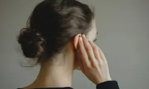 Une femme aux cheveux bruns en chignon touche son oreille, suggérant une solution naturelle comme l'huile d'amande pour les bouchons d'oreille.