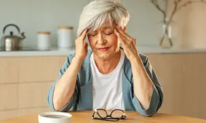 Femme âgée se tenant les tempes, souffrant de fatigue intense, vertige et palpitations. Une tasse de café et des lunettes sont sur la table.