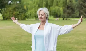 Femme d'âge mûr pratiquant le mouvement qi gong dans un parc, bras écartés, yeux fermés, en harmonie avec la nature.