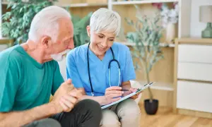Une aide-soignante discute avec un patient âgé, lui expliquant son plan de soins. Elle tient un dossier, et le patient semble attentif. La question "combien d'heures d'aide pour un gir 3" est au cœur de leur échange.