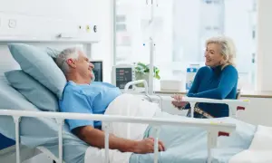 Un homme âgé, en chemise d'hôpital, sourit à une femme assise à son chevet. Une scène réconfortante d'apa et d'hospitalisation.