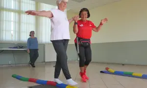 Deux seniors s'exercent sur un parcours équilibre, bras écartés pour maintenir leur stabilité. Une troisième femme observe.
