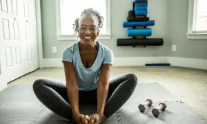Femme souriante en position de papillon sur un tapis de yoga, prête pour son **gym pour seniors 80 ans**. Haltères roses à proximité.