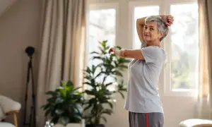 Une femme senior souriante pratique un assouplissement du dos, bras levés, dans un salon lumineux.