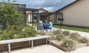 Trois femmes assises sur un banc dans un jardin fleuri, devant des maisons modernes. Un lieu de vie paisible pour un village Alzheimer à Dax.