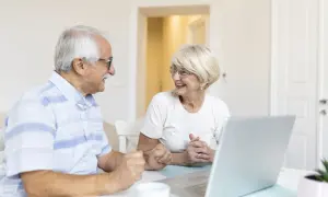 Couple âgé souriant devant un ordinateur, discutant de leur compte bancaire sous curatelle renforcée.