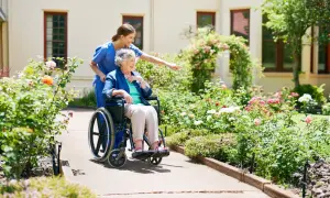 Une aide-soignante pousse une dame âgée en fauteuil roulant dans un jardin fleuri. Une scène paisible, loin des préoccupations de placement en EHPAD sans consentement.