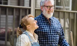 Couple souriant devant une petite maison pour personne âgée. L'homme porte une chemise à carreaux, la femme une salopette en jean.