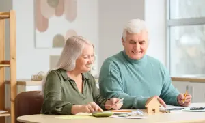 Couple âgé planifiant leur budget pour l'entrée en EHPAD et la résidence principale, avec une calculatrice et une petite maison en bois.