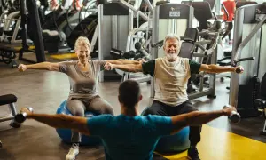 Un couple de retraités souriants fait un exercice swiss ball avec des haltères, guidé par un coach.