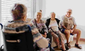 Quatre personnes âgées discutent sur un canapé. L'une est en fauteuil roulant. Le tarif du foyer logement senior est abordé.