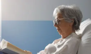 Une femme âgée, en **had et apa**, lit un livre confortablement installée dans un lit.