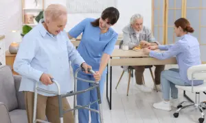 Une aide-soignante accompagne une résidente d'un EHPAD Alzheimer avec un déambulateur. Au fond, d'autres résidents sont à table.