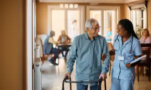 Une aide-soignante soutient un homme âgé marchant avec un déambulateur dans un foyer logement.