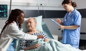 Soins attentifs pour une **hospitalisation à domicile cancer phase terminale**. Une infirmière écoute le cœur d'un patient âgé, une autre note des informations.