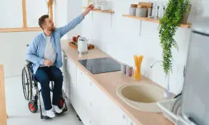 Un homme en fauteuil roulant attrape un objet sur une étagère dans une cuisine adaptée. Un exemple de priorité handicap logement social.