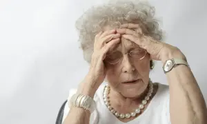 Une femme âgée, aux cheveux gris bouclés, se tient la tête, le regard perdu. Elle porte des lunettes, un collier de perles et un bracelet. Son expression suggère une détresse, peut-être liée à un stade avancé de la maladie d'Alzheimer.