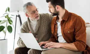 Un homme âgé explique à un plus jeune pourquoi doit on rembourser l'APA, en regardant un ordinateur portable.