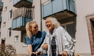 Une aide-soignante soutient une femme âgée riant aux éclats, une scène de vie chaleureuse qui pourrait inspirer une alternative maison de retraite.