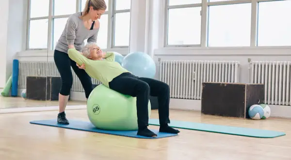 Une jeune femme aide une senior à faire des exercices sur un ballon de gym. Le **gym dos senior** est une activité bénéfique.