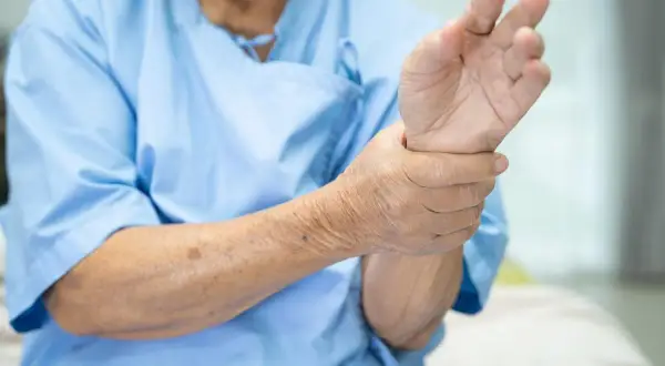 Main d'une personne âgée en blouse bleue, tenant son poignet, peut-être un pouce qui bouge tout seul.