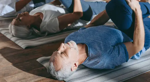 Des seniors pratiquent la gym douce sur des tapis. Un homme et une femme, les genoux pliés, se détendent.