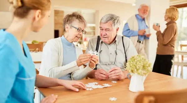 Des aînés assemblent un puzzle dans une maison de retraite gratuite, aidés par une aide soignante.