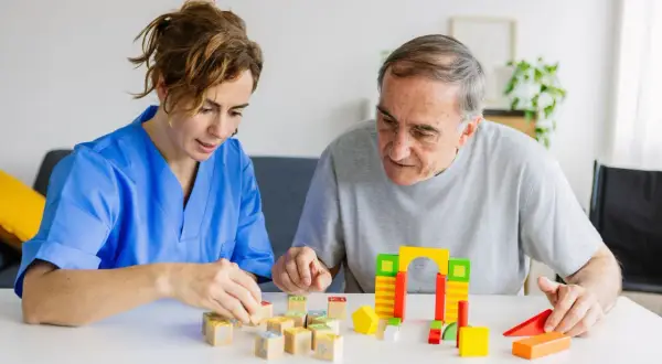 Une aide-soignante en blouse bleue joue avec des cubes en bois avec un homme âgé. Une scène de la vie quotidienne dans un établissement de soins, pas un pasa ehpad.
