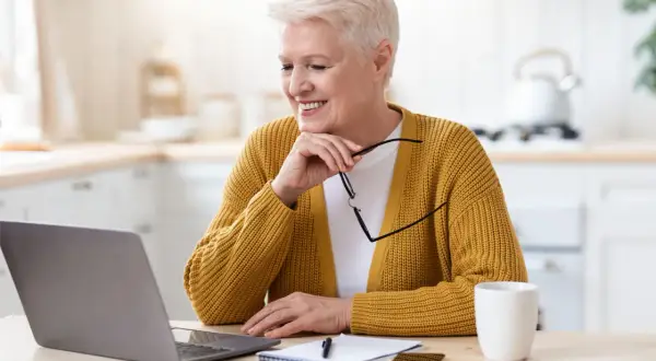Femme souriante devant son ordinateur, lunettes à la main, peut-être en train de consulter la liste des uhr en France.