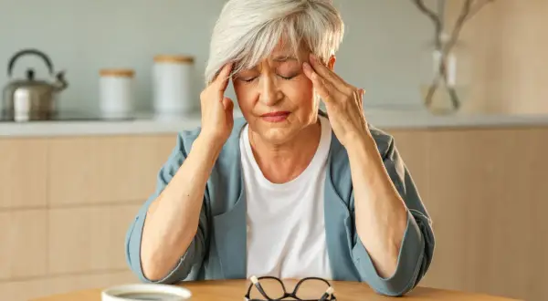 Femme âgée se tenant les tempes, souffrant de fatigue intense, vertige et palpitations. Une tasse de café et des lunettes sont sur la table.