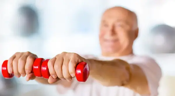 Un homme souriant, la soixantaine bien entamée, fait des exercices avec des haltères rouges. Un exemple de gym pour seniors 70 ans.