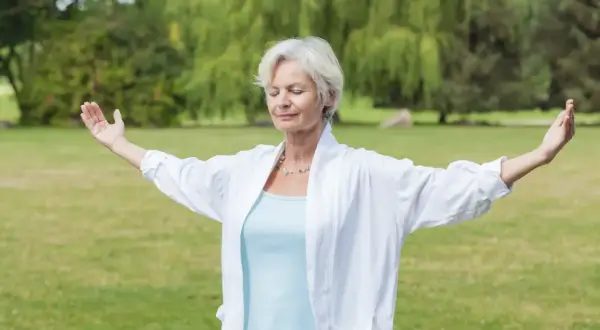 Femme d'âge mûr pratiquant le mouvement qi gong dans un parc, bras écartés, yeux fermés, en harmonie avec la nature.