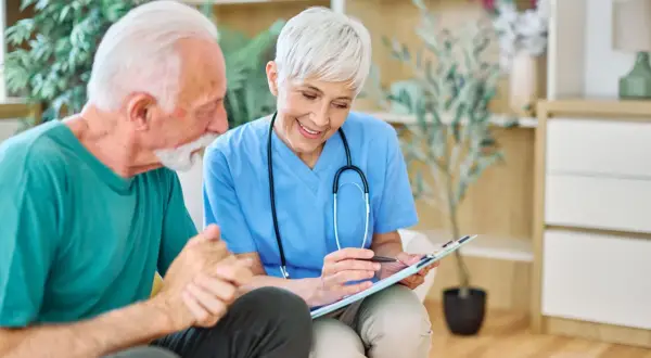 Une aide-soignante discute avec un patient âgé, lui expliquant son plan de soins. Elle tient un dossier, et le patient semble attentif. La question "combien d'heures d'aide pour un gir 3" est au cœur de leur échange.