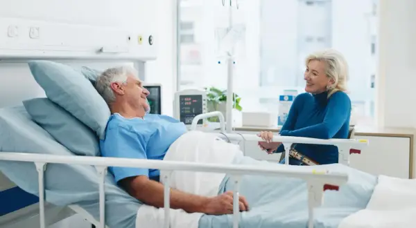 Un homme âgé, en chemise d'hôpital, sourit à une femme assise à son chevet. Une scène réconfortante d'apa et d'hospitalisation.