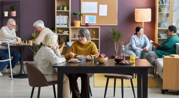 Des résidents profitent d'un repas convivial dans une maison partagée senior. L'un d'eux mange une soupe, tandis que d'autres jouent aux échecs ou discutent.