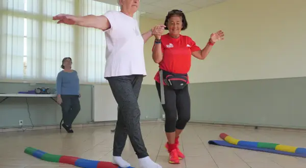 Deux seniors s'exercent sur un parcours équilibre, bras écartés pour maintenir leur stabilité. Une troisième femme observe.