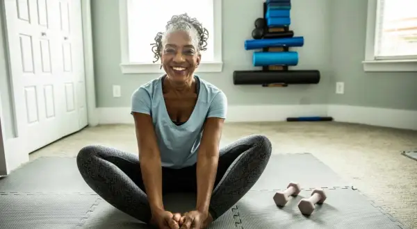 Femme souriante en position de papillon sur un tapis de yoga, prête pour son **gym pour seniors 80 ans**. Haltères roses à proximité.