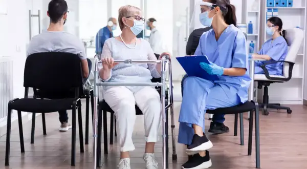Une patiente âgée avec un déambulateur discute avec une infirmière masquée dans une salle d'attente d'un usld hôpital.