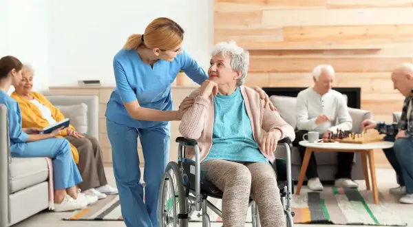 Une aide-soignante soutient une femme en fauteuil roulant, tandis que d'autres résidents jouent aux échecs. L'image évoque le soin et la transmission, comme une succession de moments de vie.