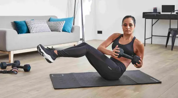 Femme faisant des torsions russes avec haltère, un exercice de gym facile pour débutant, dans un salon moderne.