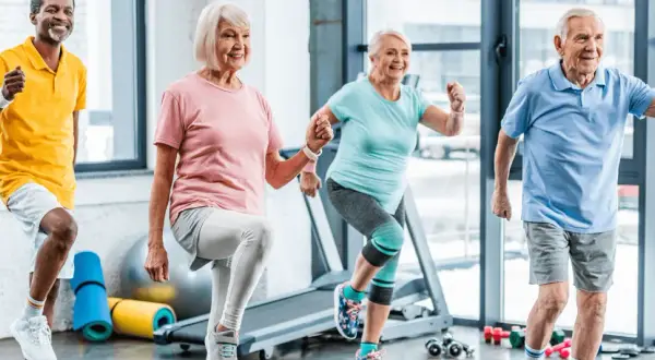 Quatre seniors souriants participent à un cours de gym, faisant des exercices dynamiques dans une salle lumineuse.