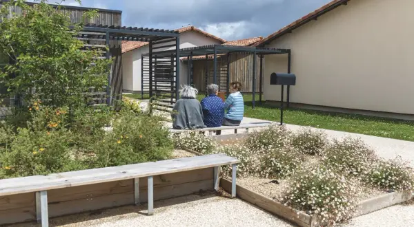 Trois femmes assises sur un banc dans un jardin fleuri, devant des maisons modernes. Un lieu de vie paisible pour un village Alzheimer à Dax.