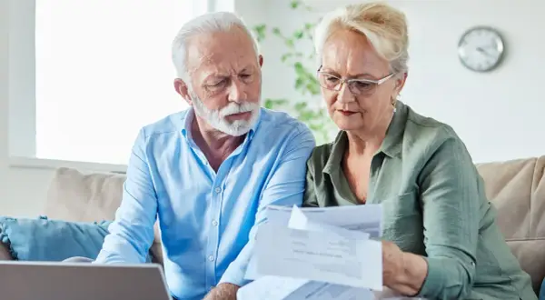 Couple âgé examinant des papiers, peut-être pour une aide EHPAD GIR 2. Ils sont sur un canapé avec un ordinateur portable.