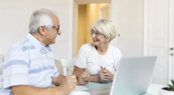 Couple âgé souriant devant un ordinateur, discutant de leur compte bancaire sous curatelle renforcée.