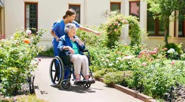 Une aide-soignante pousse une dame âgée en fauteuil roulant dans un jardin fleuri. Une scène paisible, loin des préoccupations de placement en EHPAD sans consentement.