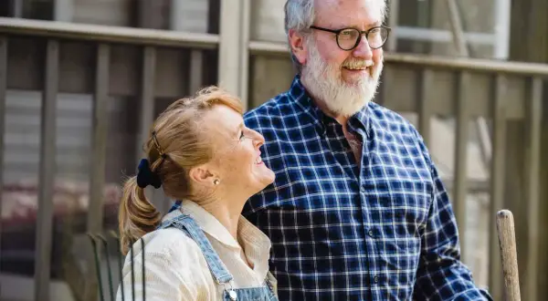 Couple souriant devant une petite maison pour personne âgée. L'homme porte une chemise à carreaux, la femme une salopette en jean.