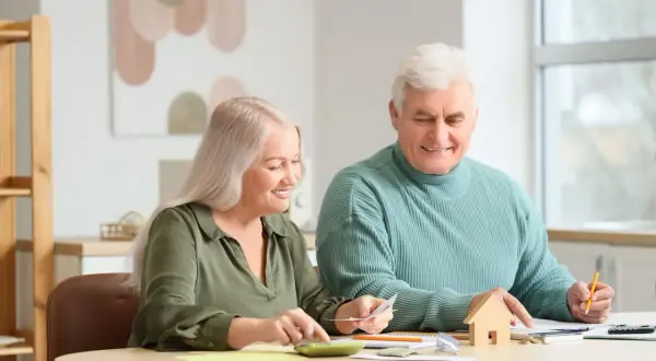 Couple âgé planifiant leur budget pour l'entrée en EHPAD et la résidence principale, avec une calculatrice et une petite maison en bois.