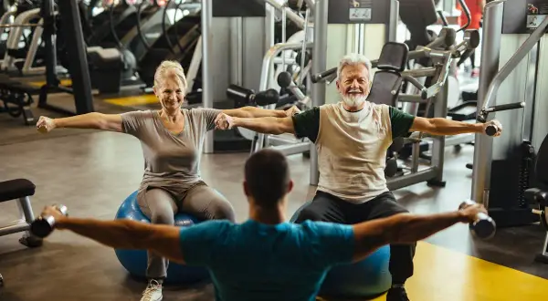 Un couple de retraités souriants fait un exercice swiss ball avec des haltères, guidé par un coach.